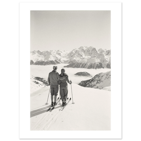 Ski couple resting on a snowy alpine summit with skis planted, looking over a range of peaks, 1932 style black and white photo print